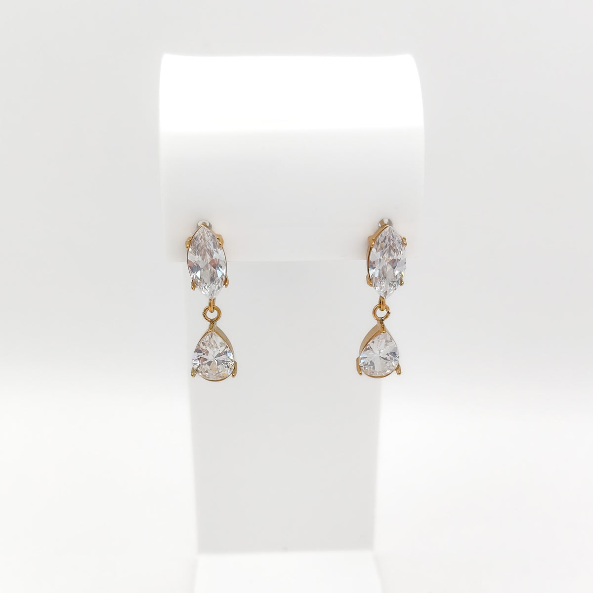 A pair of gold drop earrings with marquise-cut clear gemstones on the top and pear-cut clear gemstones on the bottom, displayed on a white jewelry stand against a white background.