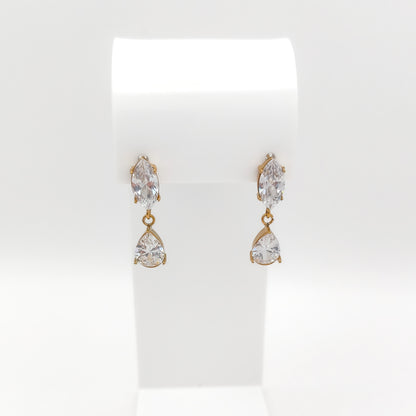A pair of gold drop earrings with marquise-cut clear gemstones on the top and pear-cut clear gemstones on the bottom, displayed on a white jewelry stand against a white background.