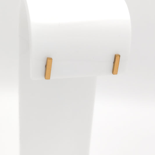 Detailed close-up of a pair of gold-plated stainless steel minimalist vertical bar stud earrings with a high-shine polished finish, displayed on a white stand.