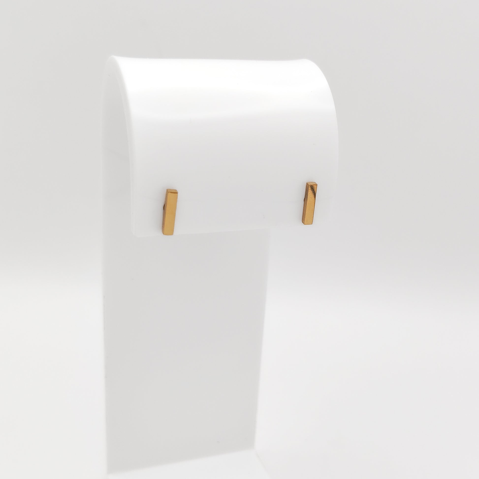 A pair of gold-plated stainless steel minimalist vertical bar stud earrings with a high-shine polished finish, displayed on a white stand.