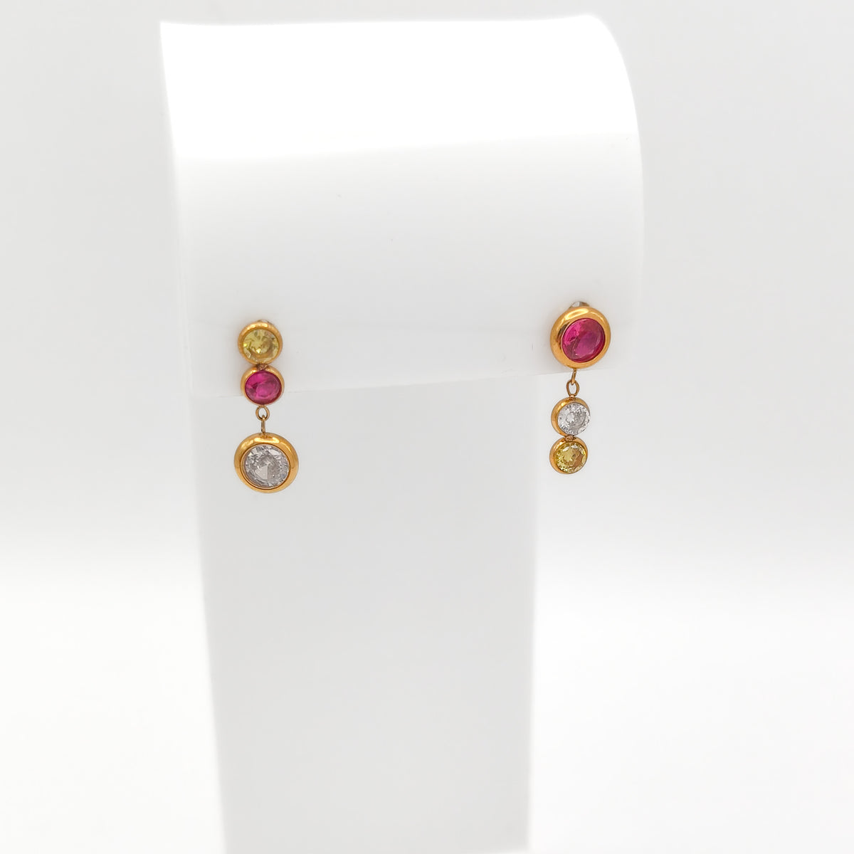 Gold-plated stainless steel mismatch bezel-set drop earrings with pink, yellow, and clear round cubic zirconia stones on a white stand.