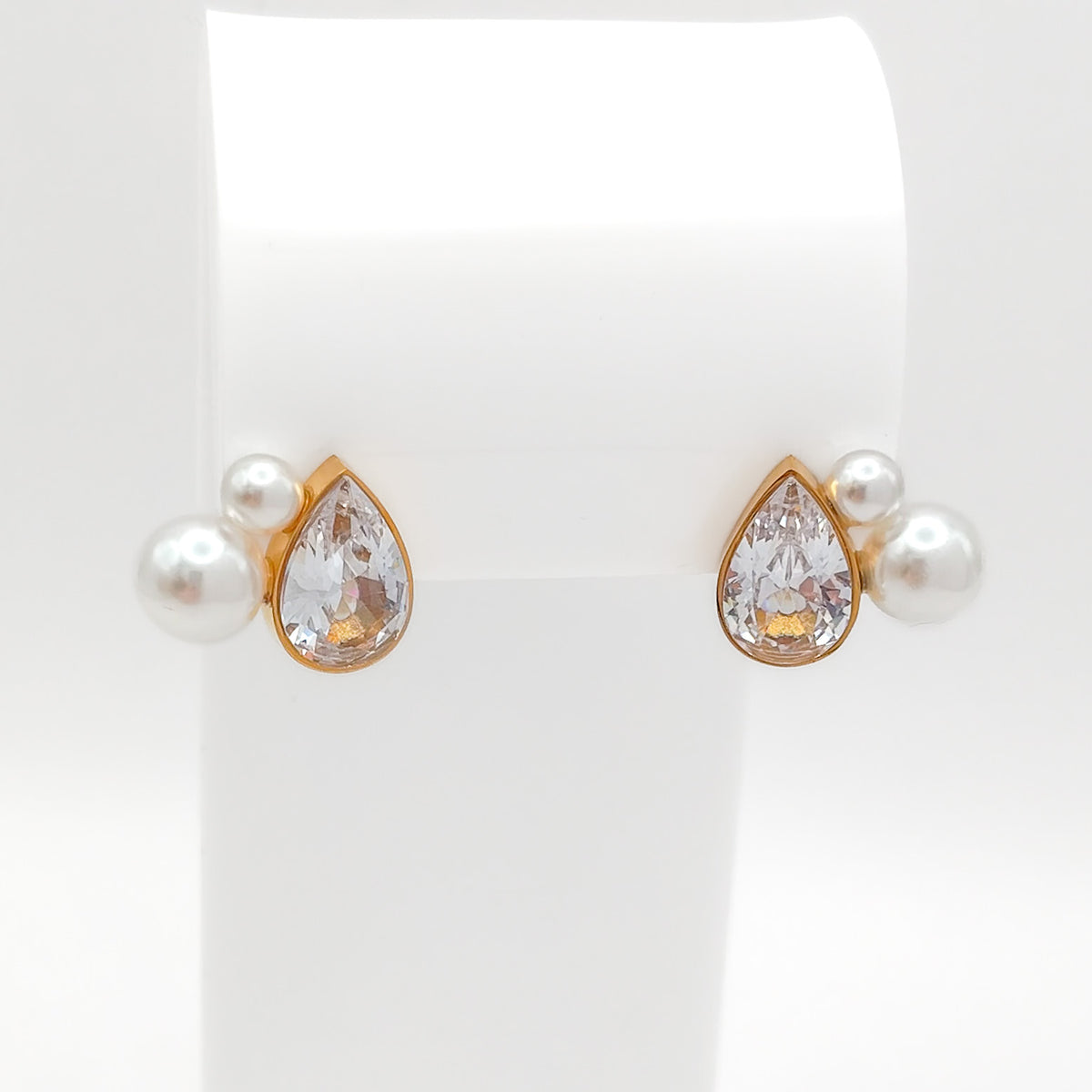 Front view of gold-plated stainless steel stud earrings showcasing a bezel-set pear-cut CZ stone flanked by small white pearls on a white stand.