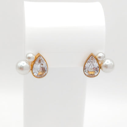 Front view of gold-plated stainless steel stud earrings showcasing a bezel-set pear-cut CZ stone flanked by small white pearls on a white stand.