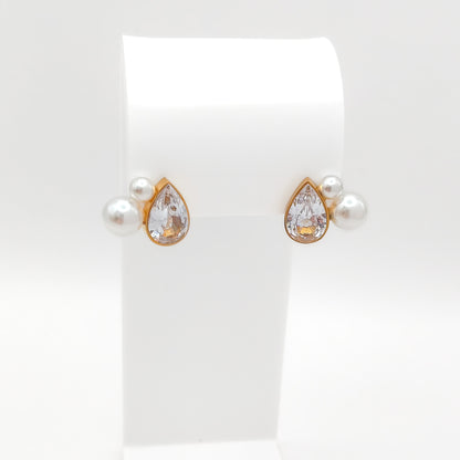 A pair of gold-plated stainless steel cluster stud earrings featuring a bezel-set teardrop cubic zirconia stone accented by two faux pearls, displayed on a white stand.