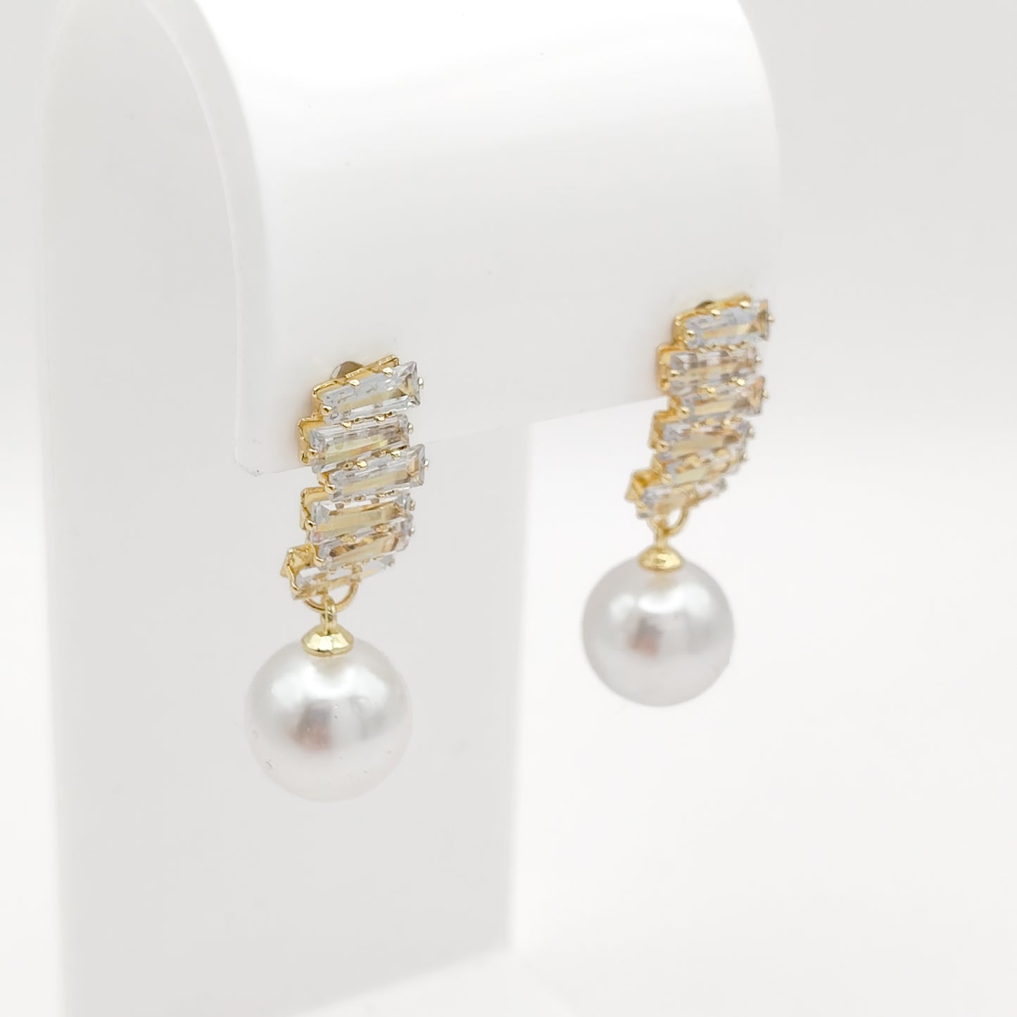 A pair of gold drop earrings with a textured, crystal-encrusted top and a dangling white pearl, displayed on a white jewelry stand.