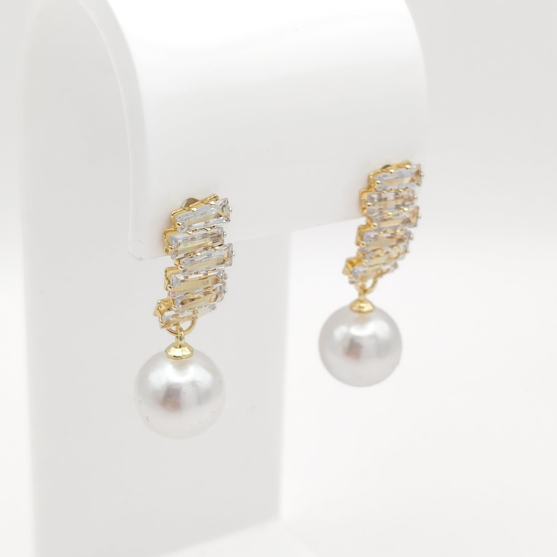 A pair of gold drop earrings with a textured, crystal-encrusted top and a dangling white pearl, displayed on a white jewelry stand.