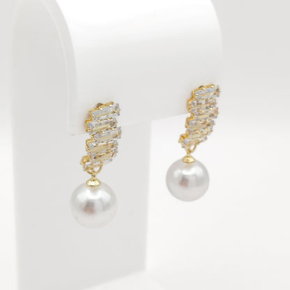 A pair of gold drop earrings with a textured, crystal-encrusted top and a dangling white pearl, displayed on a white jewelry stand.