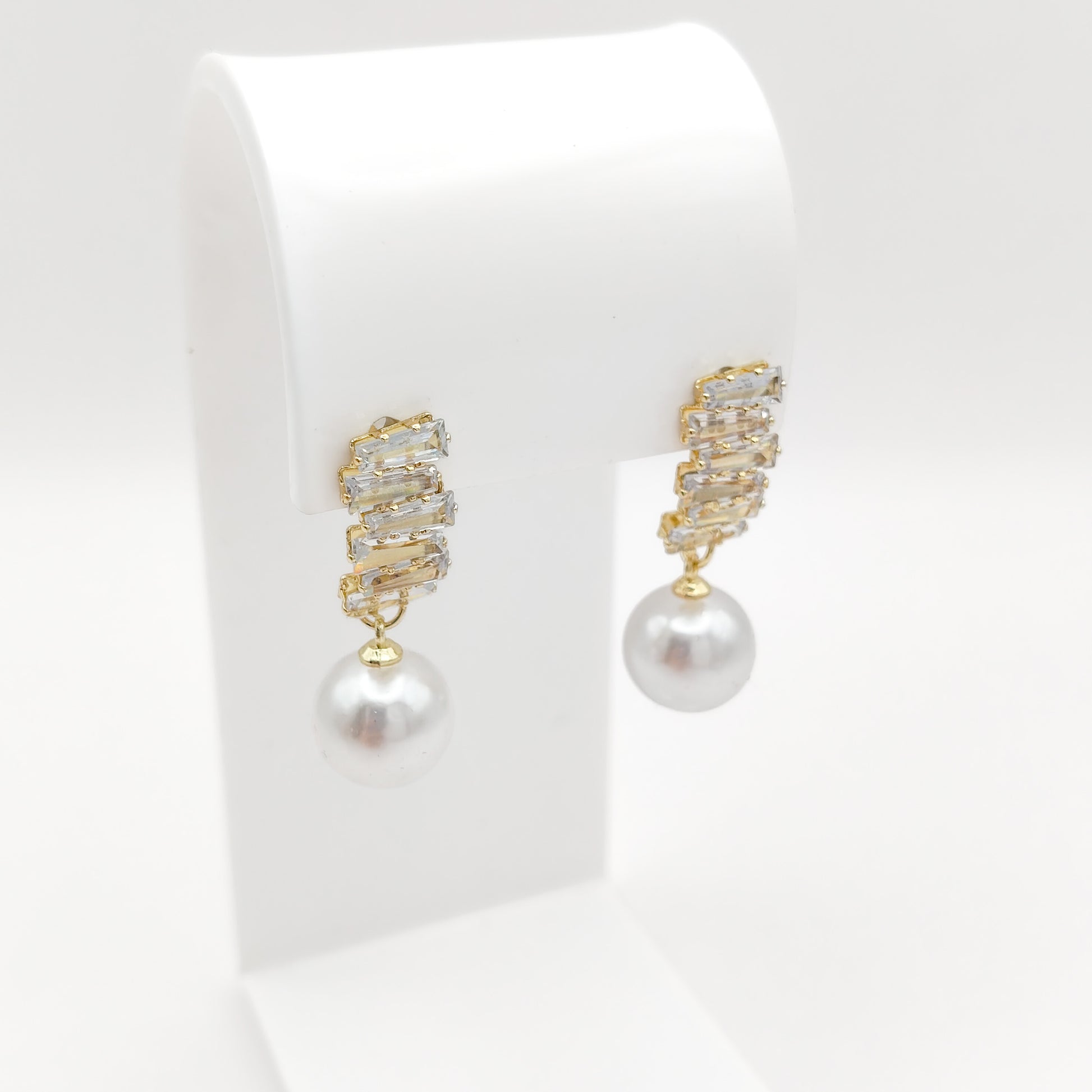 A pair of gold drop earrings with a textured, rhinestone-encrusted top and a single white pearl dangling below, displayed on a white jewelry stand against a white background.