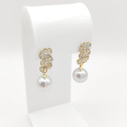 A pair of gold drop earrings with a textured, rhinestone-encrusted top and a single white pearl dangling below, displayed on a white jewelry stand against a white background.