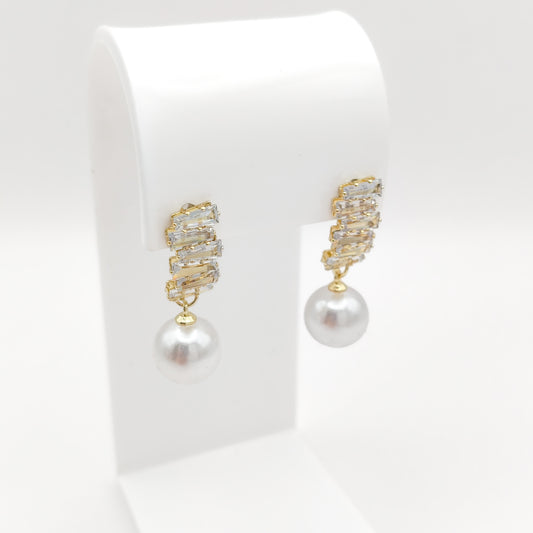 A pair of gold drop earrings with a textured, rhinestone-encrusted top and a single white pearl dangling below, displayed on a white jewelry stand against a white background.