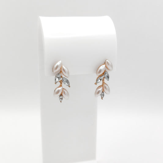 A pair of rose gold leaf drop earrings with white faux pearls and clear marquise-cut crystals, displayed on a white curved stand against a light gray background.