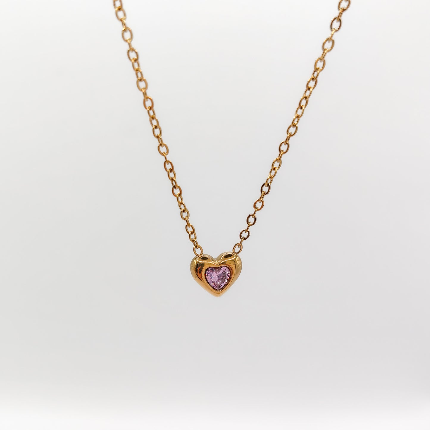 Delicate gold-plated stainless steel chain necklace with a petite heart pendant featuring a sparkling pink cubic zirconia stone, showcased on a white display stand.