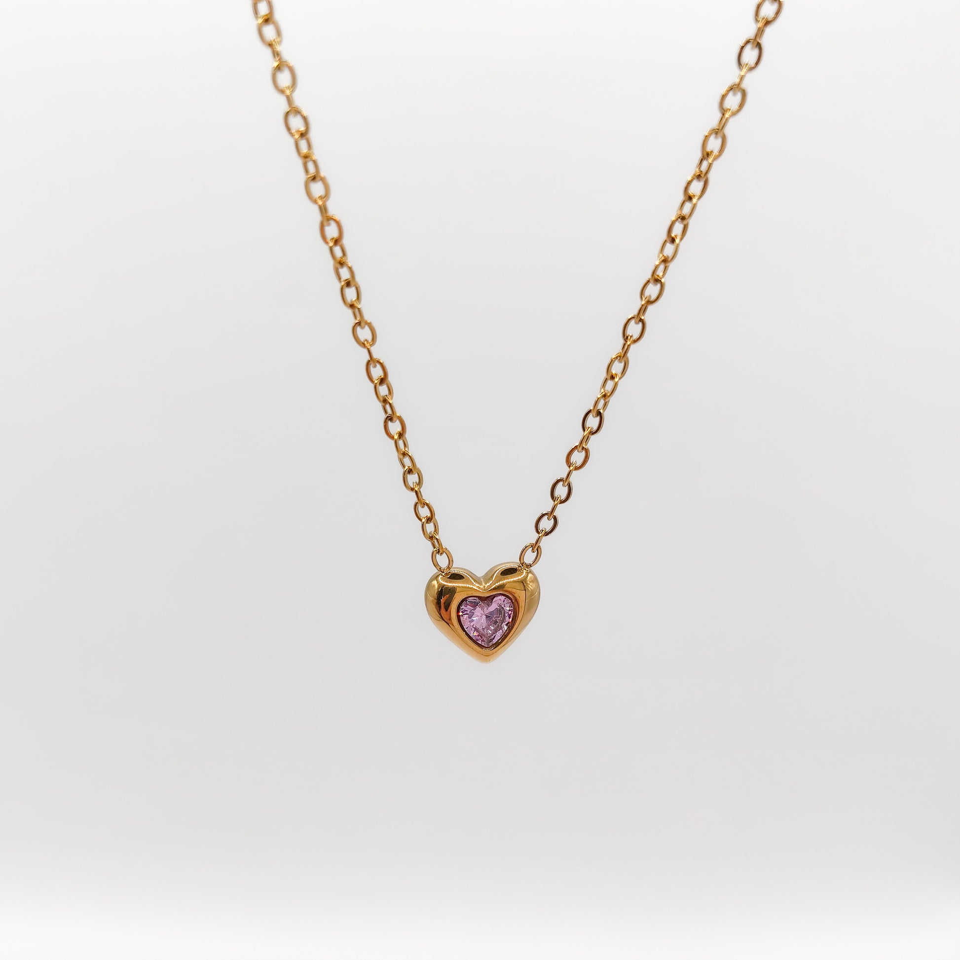 Delicate gold-plated stainless steel chain necklace with a petite heart pendant featuring a sparkling pink cubic zirconia stone, showcased on a white display stand.