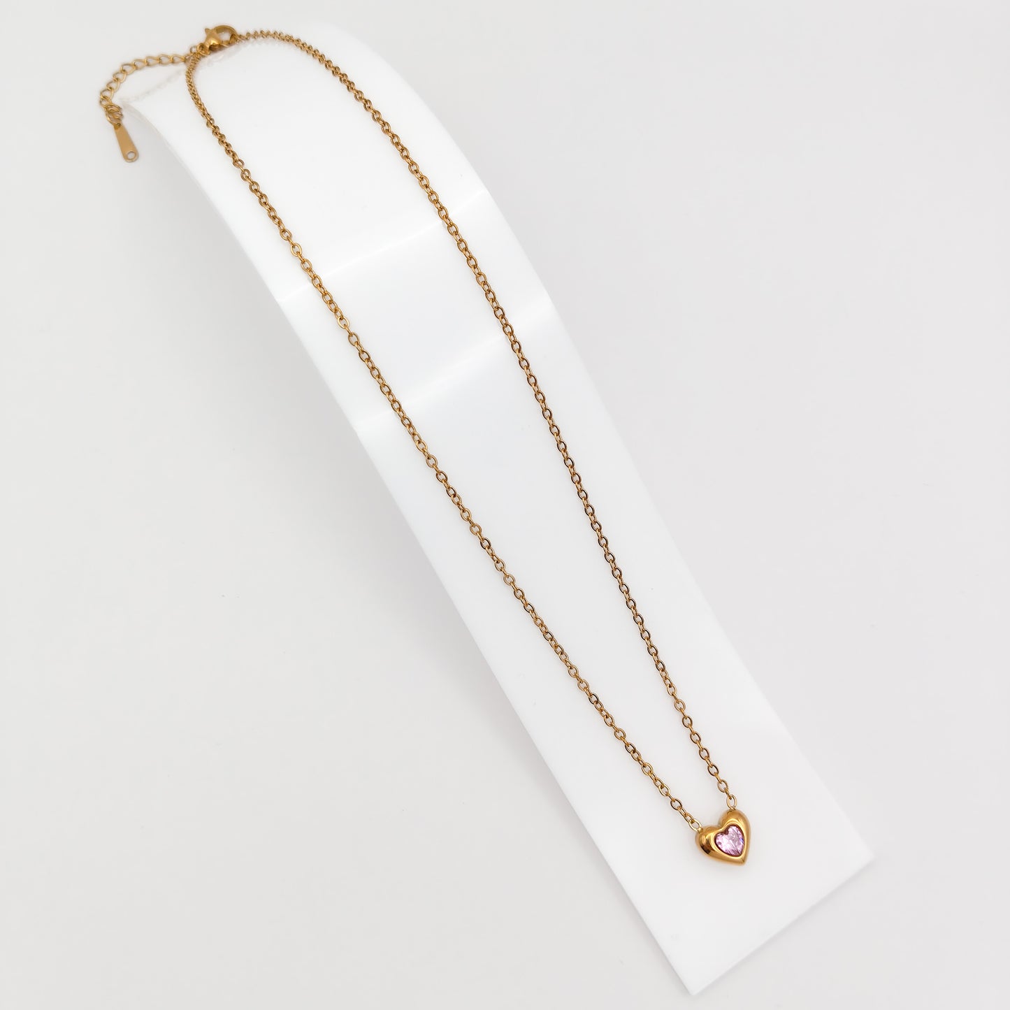 Delicate gold-plated stainless steel chain necklace with a petite heart pendant featuring a sparkling pink cubic zirconia stone, showcased on a white display stand.