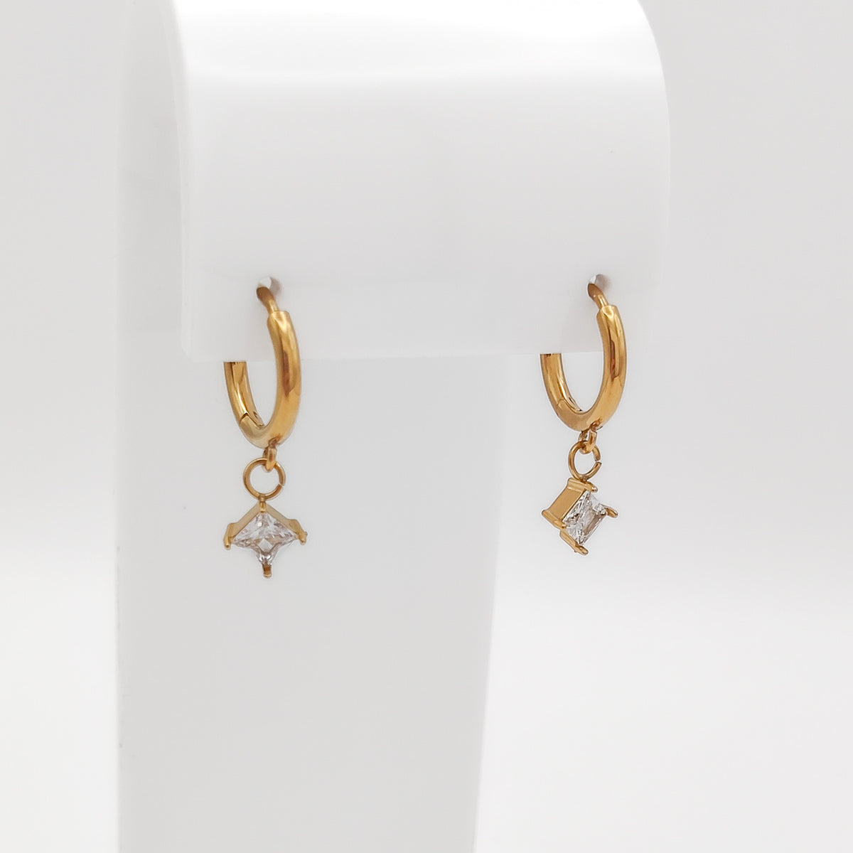 Detailed close-up of a pair of gold-plated stainless steel huggie hoop earrings with dangling square-set princess-cut clear cubic zirconia charms, displayed on a white stand.