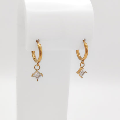 Detailed close-up of a pair of gold-plated stainless steel huggie hoop earrings with dangling square-set princess-cut clear cubic zirconia charms, displayed on a white stand.