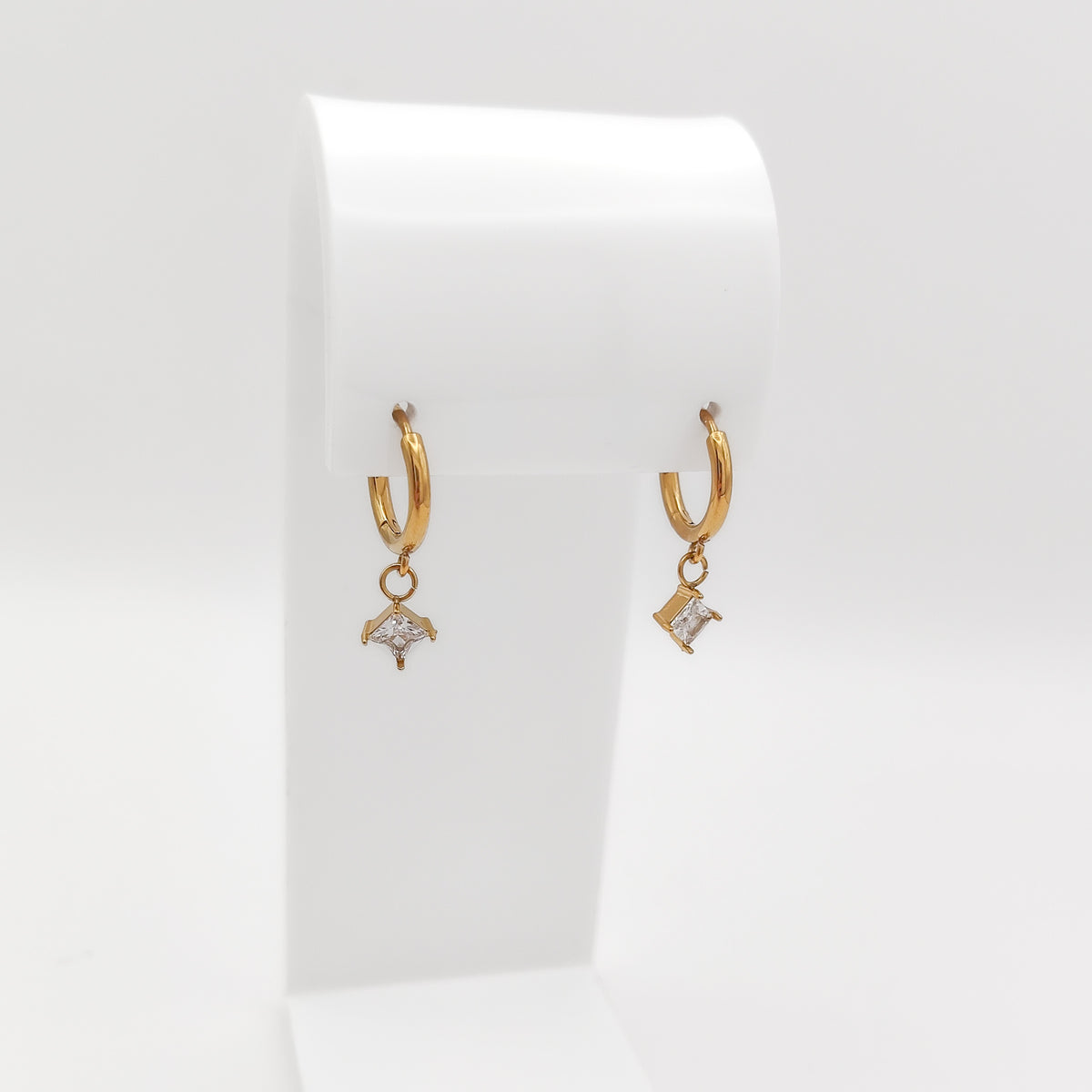 A pair of gold-plated stainless steel huggie hoop earrings with dangling square-set princess-cut clear cubic zirconia charms, displayed on a white stand.