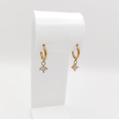 A pair of gold-plated stainless steel huggie hoop earrings with dangling square-set princess-cut clear cubic zirconia charms, displayed on a white stand.