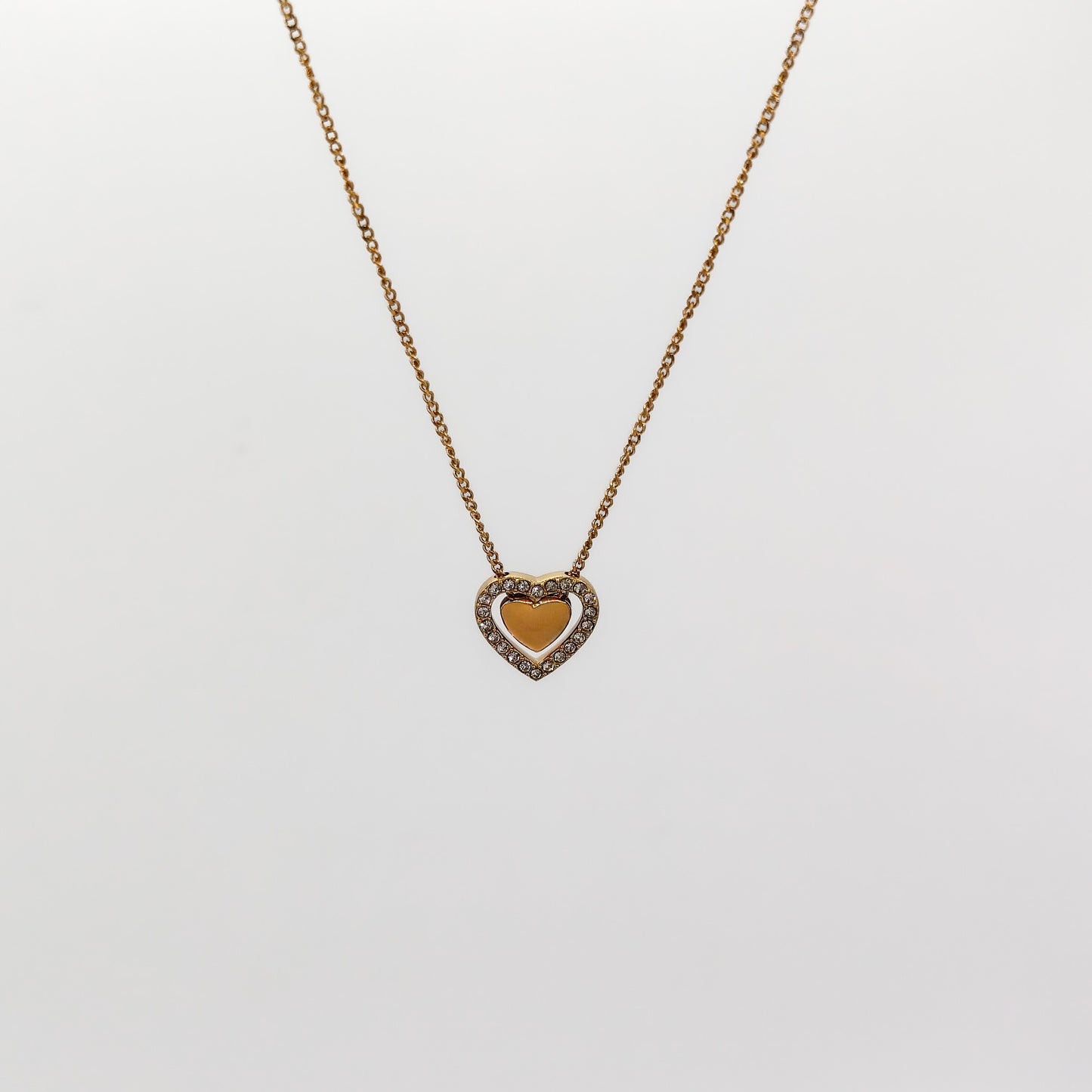 A close-up view of the gold heart-in-heart pendant, showcasing the smooth polished inner heart nestled inside a border of pavé-set clear rhinestones.