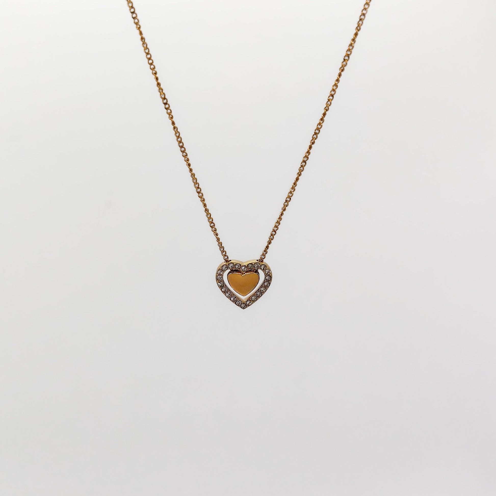 A close-up view of the gold heart-in-heart pendant, showcasing the smooth polished inner heart nestled inside a border of pavé-set clear rhinestones.
