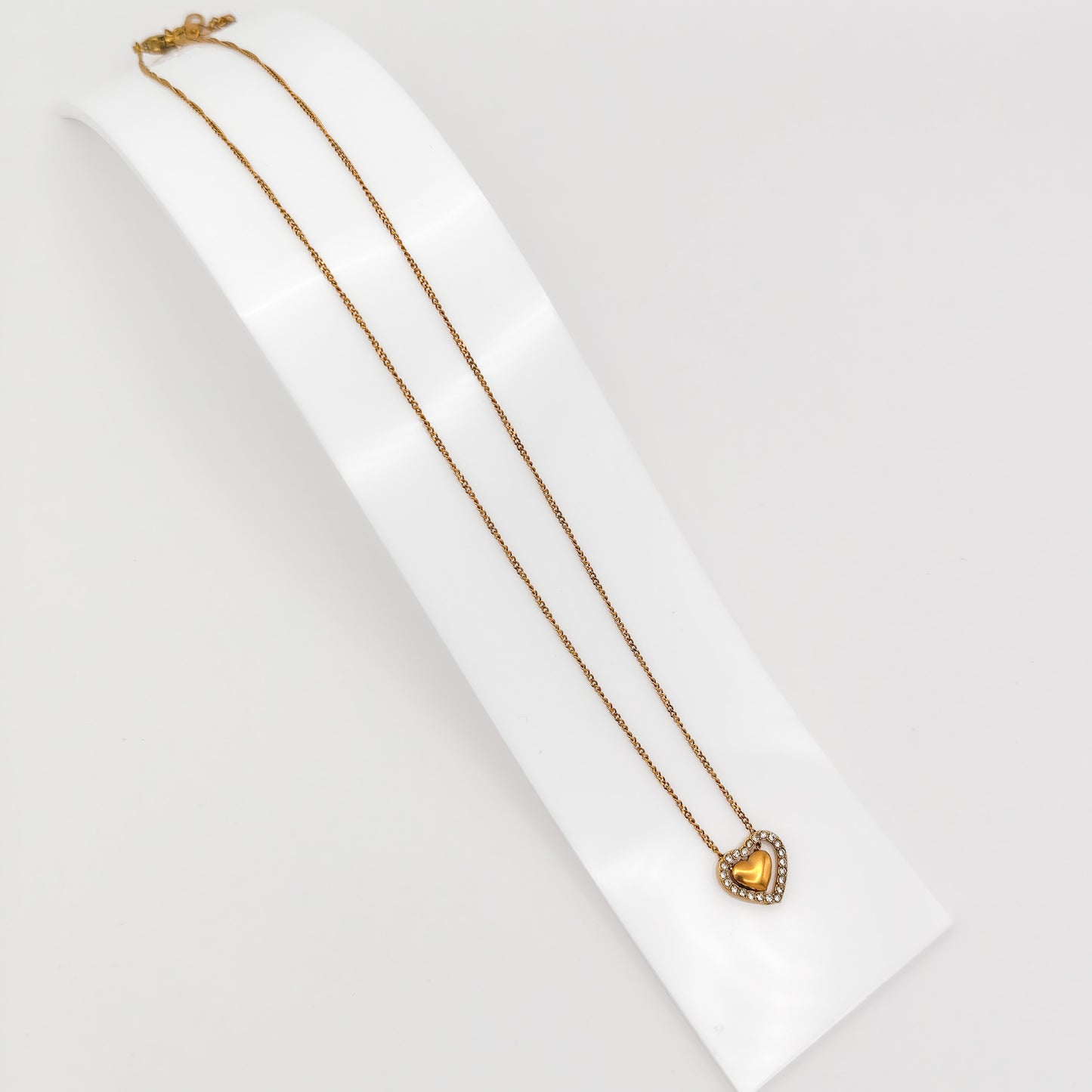 A gold-plated stainless steel princess-length chain necklace with a double heart pendant featuring a sparkling crystal rhinestone outer frame, displayed on a curved white stand.