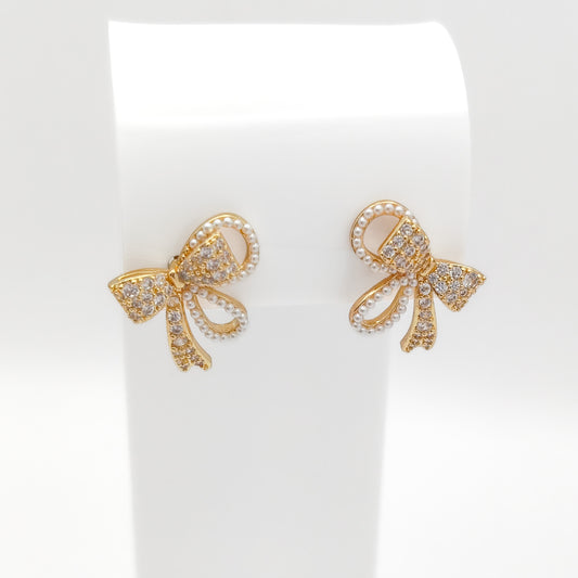 Front-facing view of gold-plated copper alloy ribbon bow stud earrings set with sparkling rhinestones and pearl trim on a white stand.