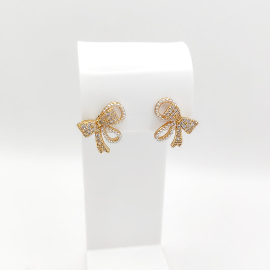 Gold-plated bow-shaped stud earrings encrusted with clear rhinestones and outlined with micro faux pearls, displayed on a white stand.