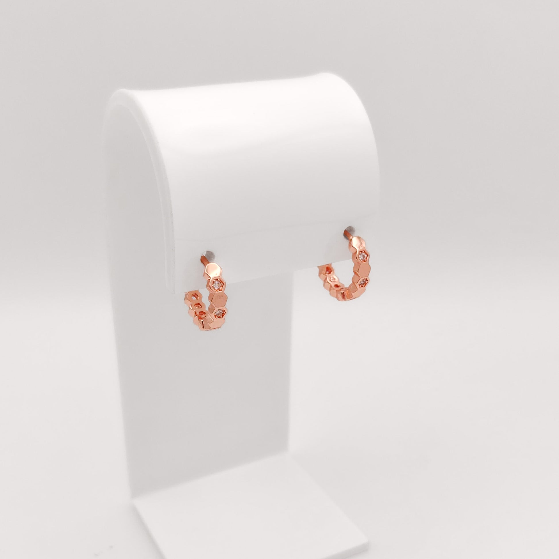 A pair of small rose gold hoop earrings with textured, beaded links, displayed on a white jewelry stand against a light gray background.