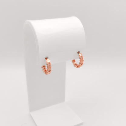 A pair of small rose gold hoop earrings with textured, beaded links, displayed on a white jewelry stand against a light gray background.