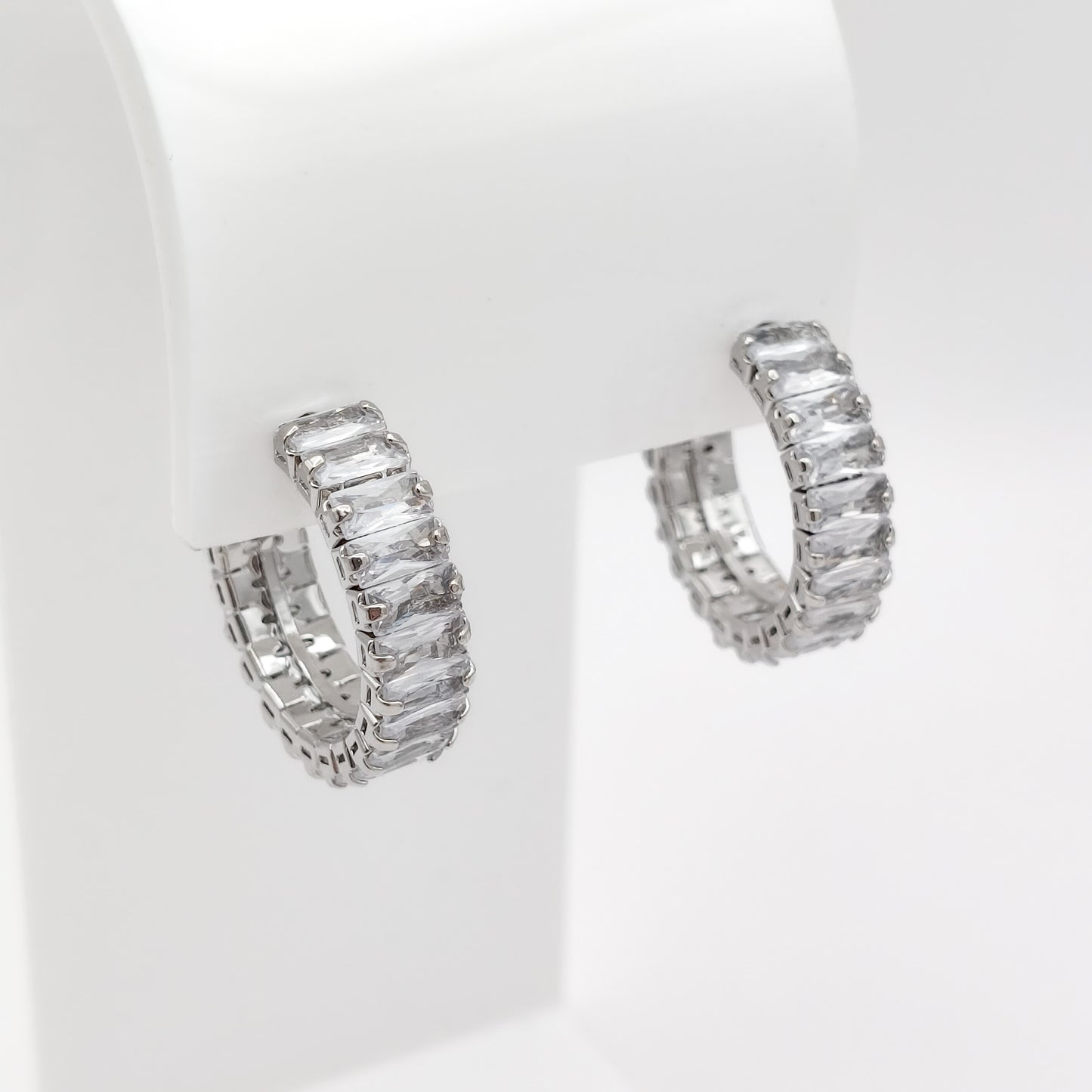A pair of 22mm silver-toned hoop earrings set with clear rectangular baguette-cut stones, displayed on a white jewelry stand against a white background.