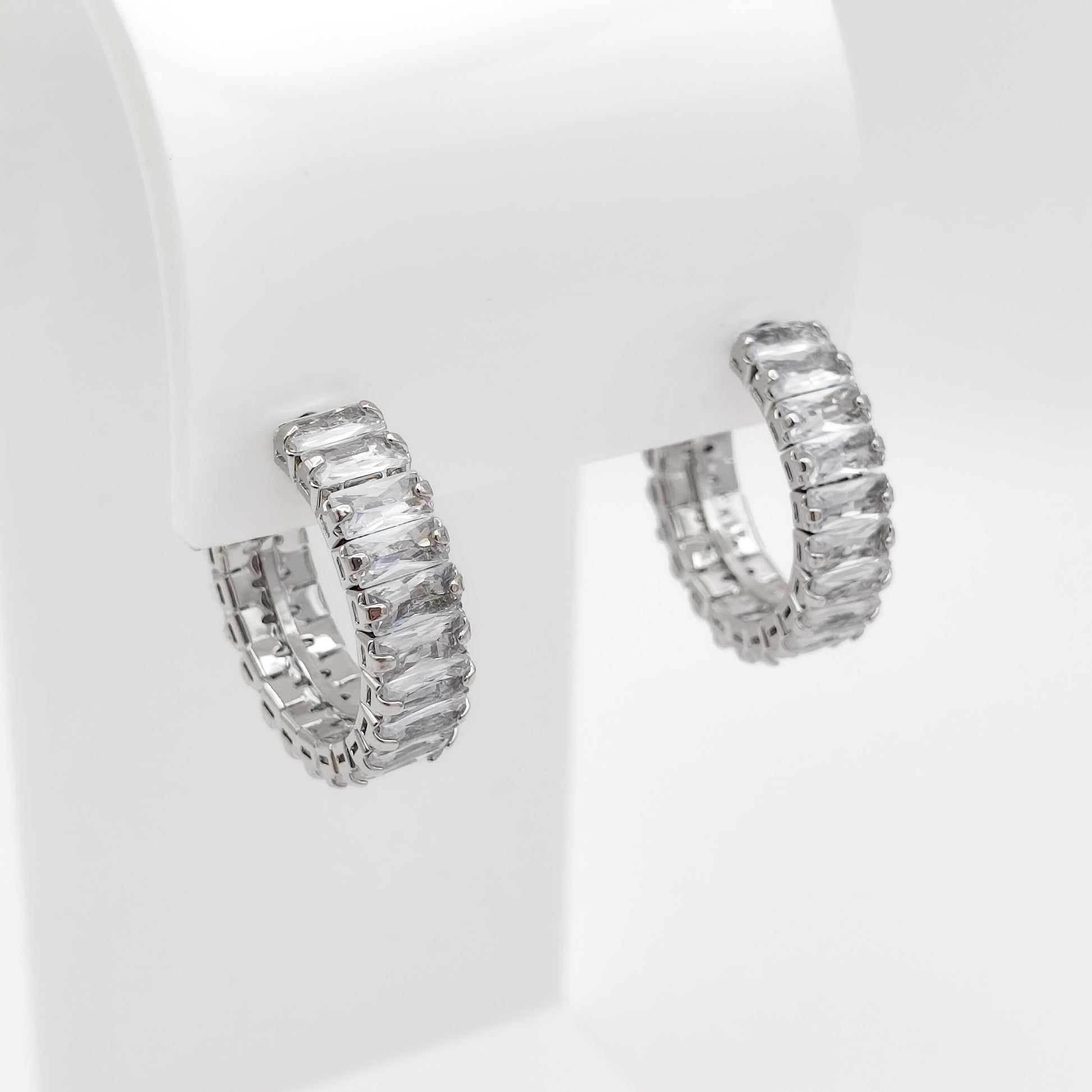 A pair of 22mm silver-toned hoop earrings set with clear rectangular baguette-cut stones, displayed on a white jewelry stand against a white background.