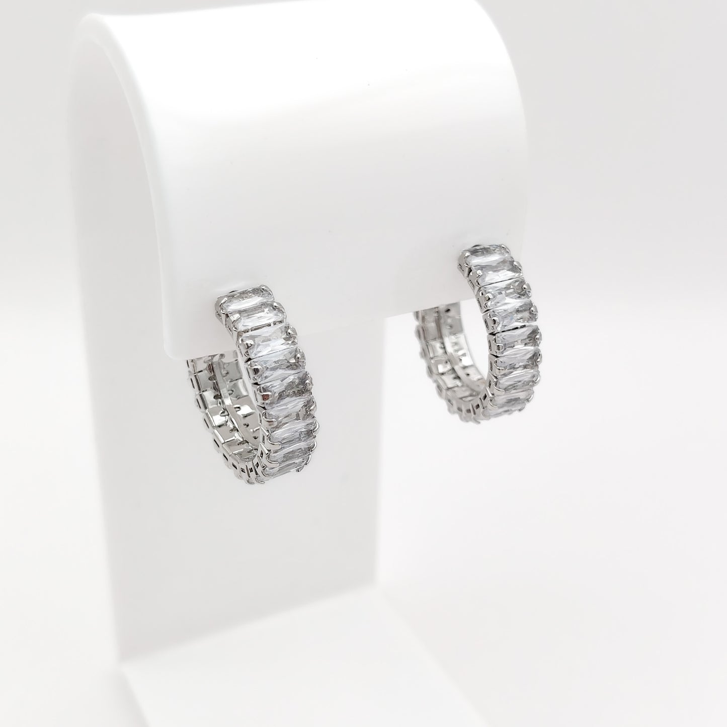 A pair of 22mm silver-plated half-hoop earrings paved with rectangular clear baguette-cut stones, displayed on a white jewelry stand against a white background.