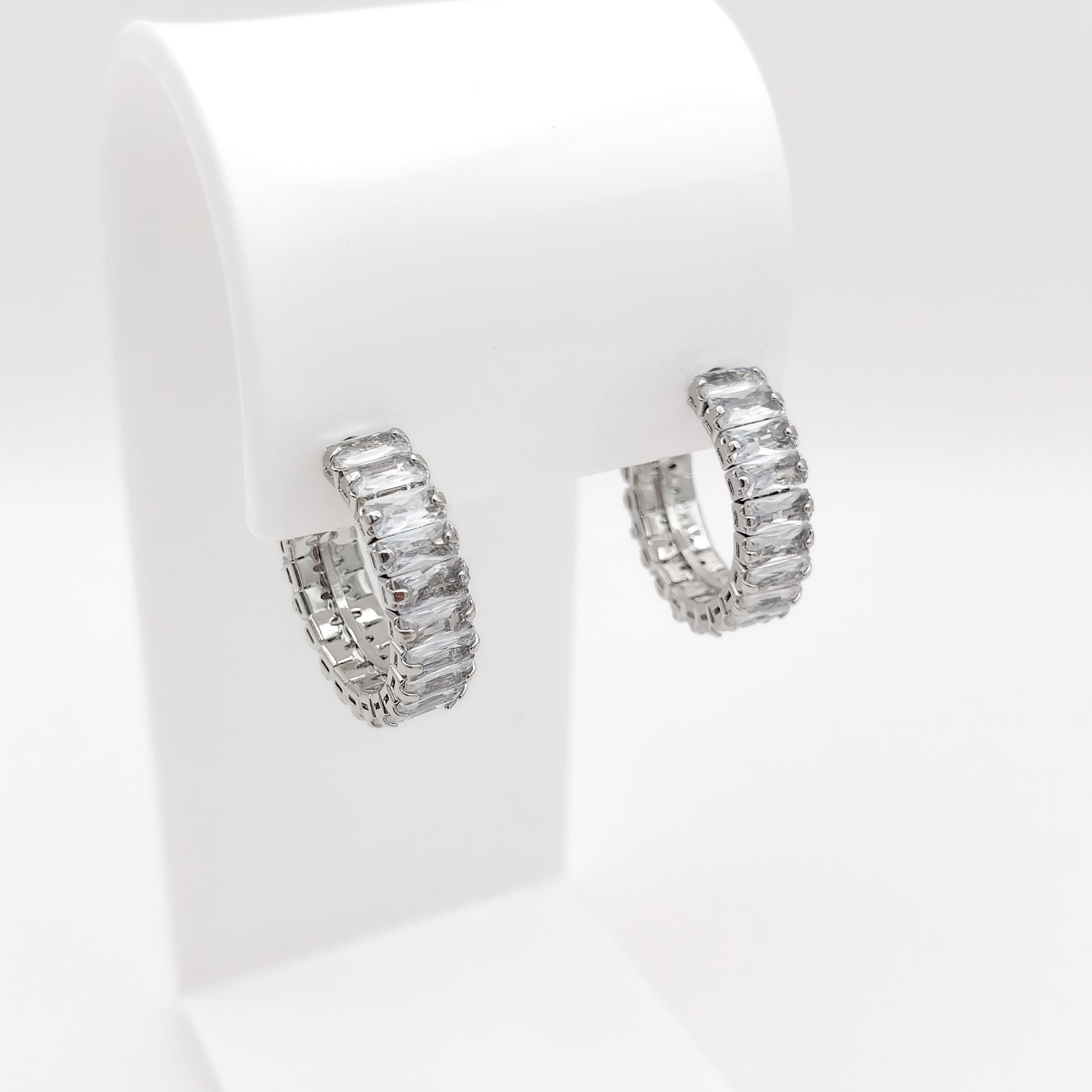 A pair of 22mm silver-plated half-hoop earrings paved with rectangular clear baguette-cut stones, displayed on a white jewelry stand against a white background.