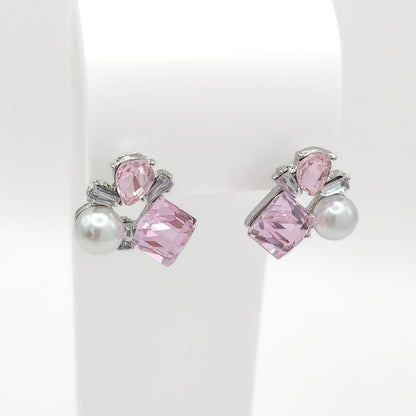 A pair of silver-toned stud earrings featuring a large faceted pink crystal, a smaller teardrop pink crystal, and a white pearl, displayed on a white jewelry stand.