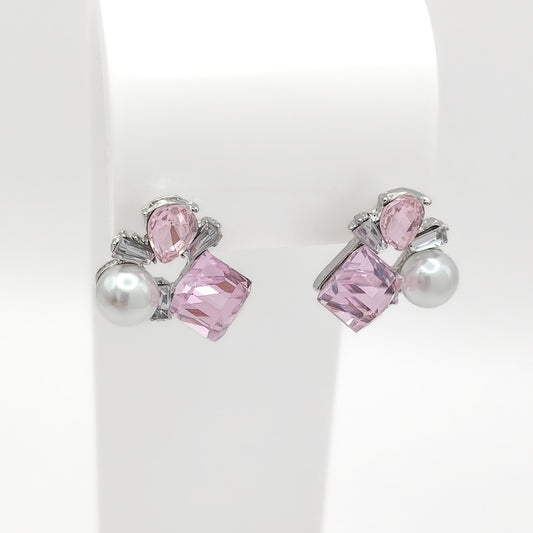 A pair of silver-toned stud earrings featuring a large faceted pink crystal, a smaller teardrop pink crystal, and a white pearl, displayed on a white jewelry stand.