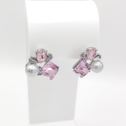 A pair of silver stud earrings with pink faceted gemstones and white faux pearls, displayed on a white jewelry stand.