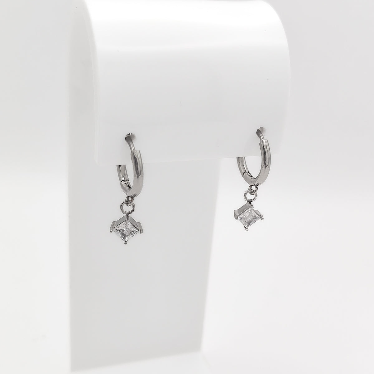 Detailed close-up image of a pair of silver-plated stainless steel huggie hoop earrings featuring dangling square princess-cut clear cubic zirconia charms, displayed on a white stand.