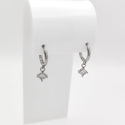 Detailed close-up image of a pair of silver-plated stainless steel huggie hoop earrings featuring dangling square princess-cut clear cubic zirconia charms, displayed on a white stand.