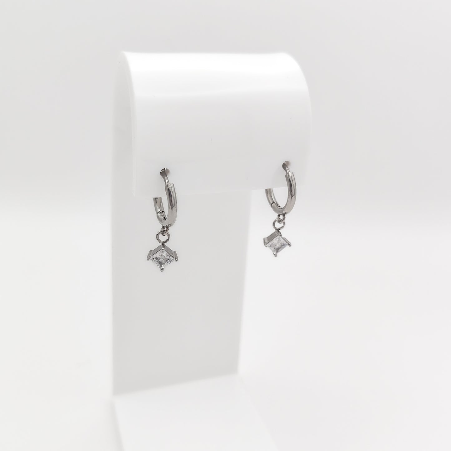 A pair of silver-plated stainless steel huggie hoop earrings featuring dangling square princess-cut clear cubic zirconia charms, displayed on a white stand.