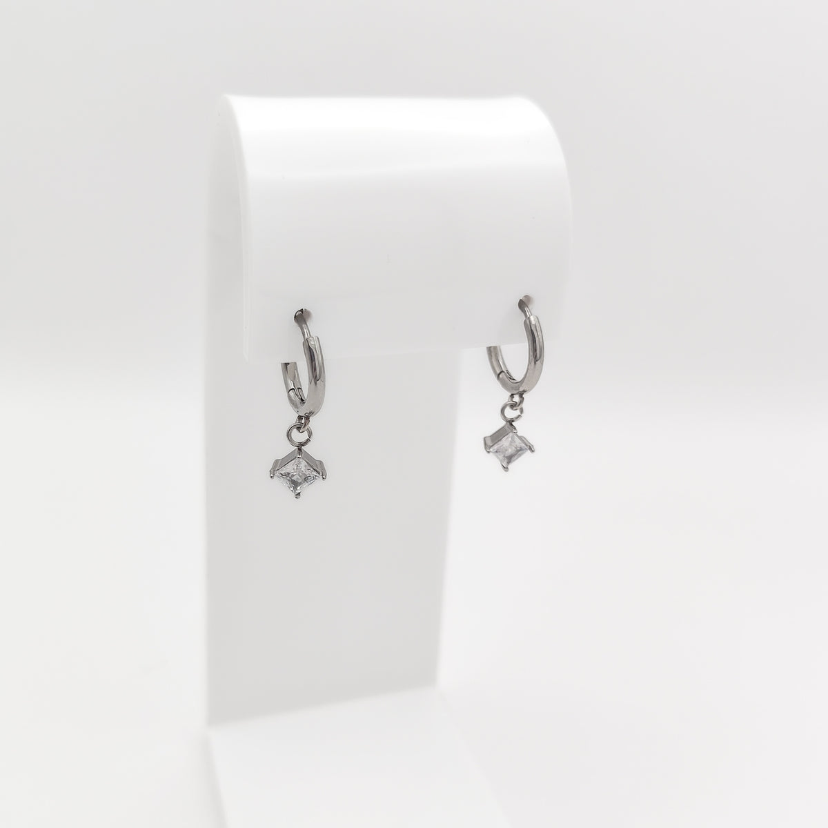 A pair of silver-plated stainless steel huggie hoop earrings featuring dangling square princess-cut clear cubic zirconia charms, displayed on a white stand.