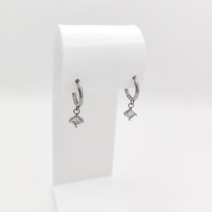 A pair of silver-plated stainless steel huggie hoop earrings featuring dangling square princess-cut clear cubic zirconia charms, displayed on a white stand.