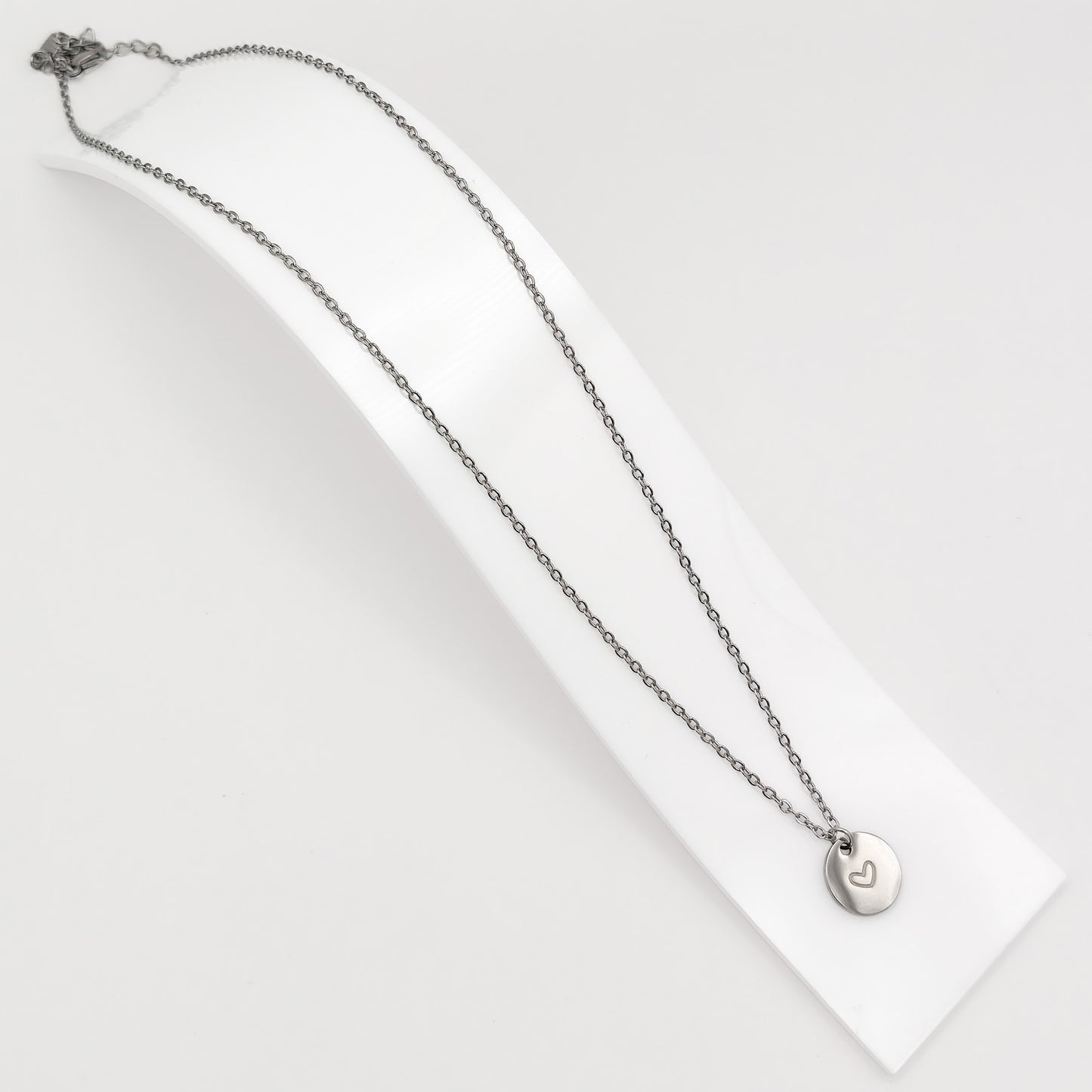 Silver plated stainless steel cable chain necklace featuring a minimalist round disc pendant with an engraved heart, displayed on a curved white stand.