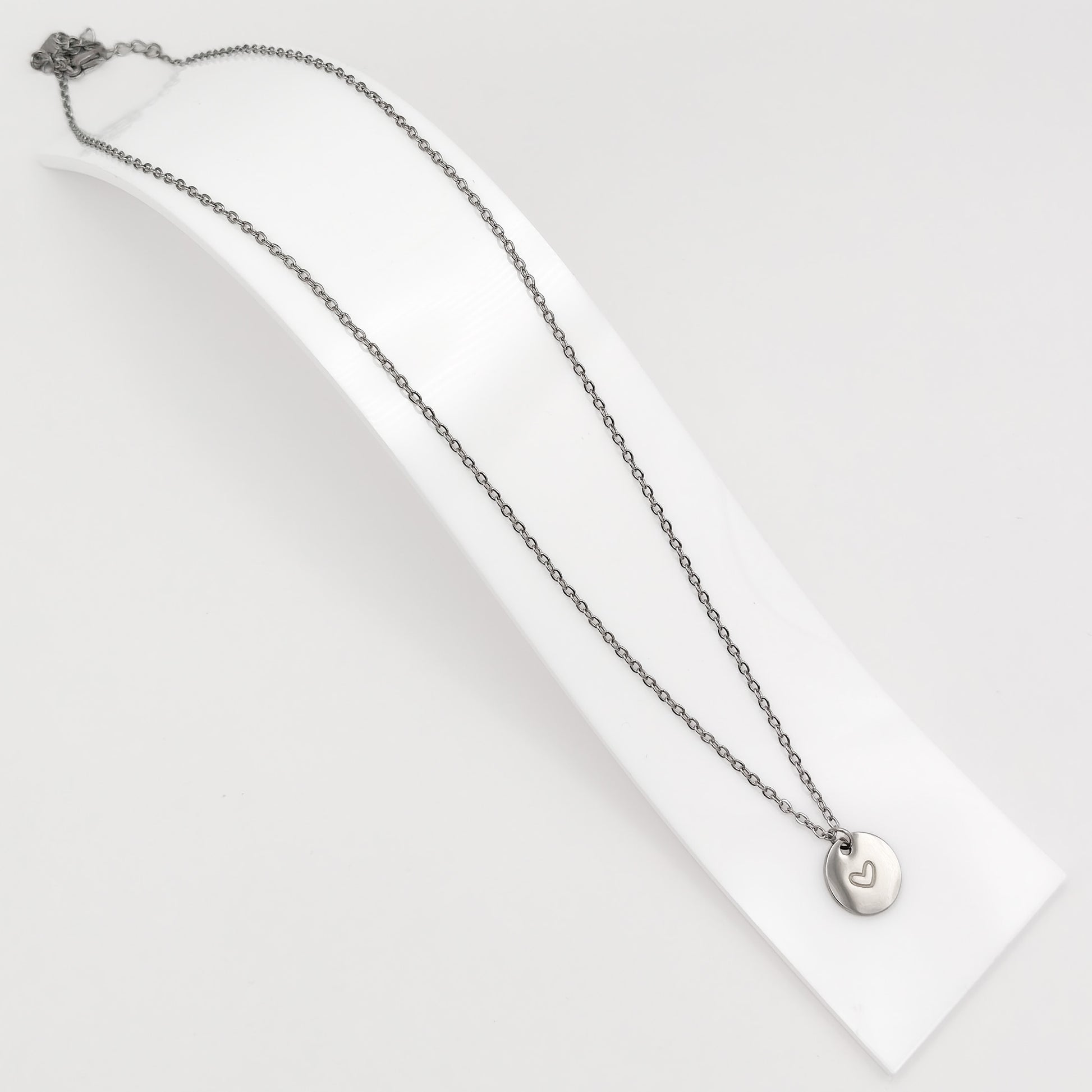Silver plated stainless steel cable chain necklace featuring a minimalist round disc pendant with an engraved heart, displayed on a curved white stand.