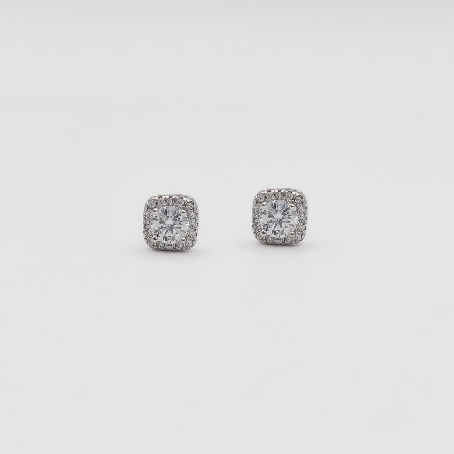 Detail image of a pair of square stud earrings with clear gemstones in a silver-toned setting, displayed on a white jewelry stand against a light gray background.
