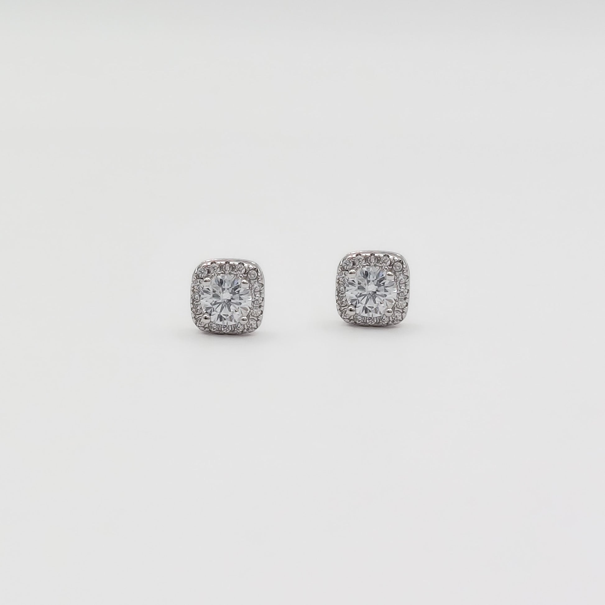 Detail image of a pair of square stud earrings with clear gemstones in a silver-toned setting, displayed on a white jewelry stand against a light gray background.