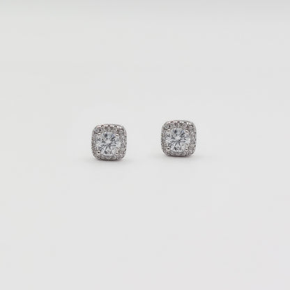 Detail image of a pair of square stud earrings with clear gemstones in a silver-toned setting, displayed on a white jewelry stand against a light gray background.