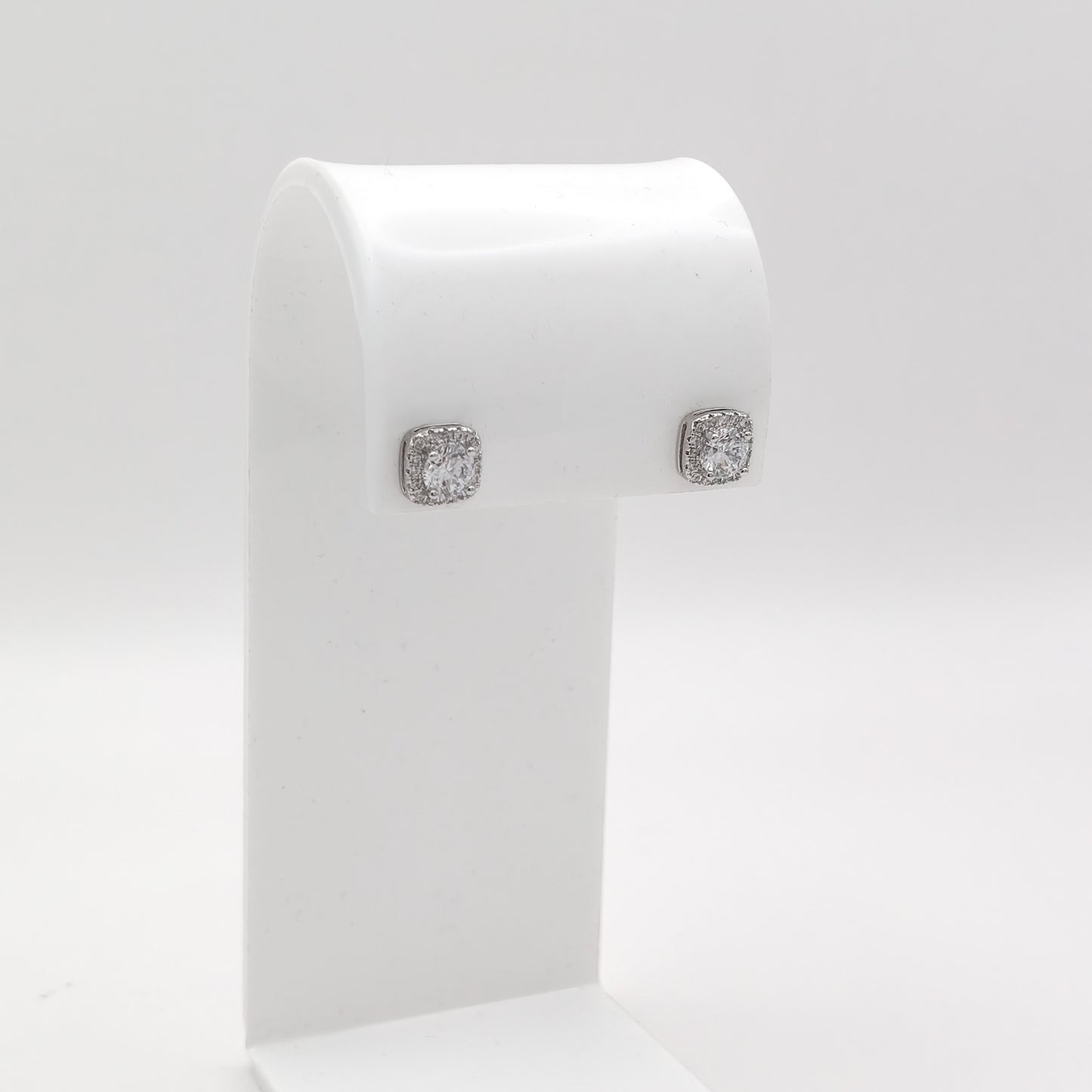 A pair of square stud earrings with clear gemstones in a silver-toned setting, displayed on a white jewelry stand against a light gray background.