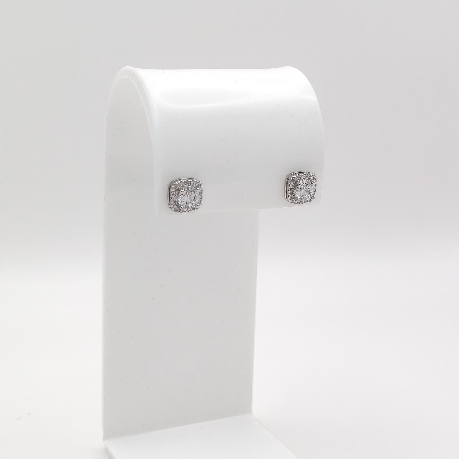 A pair of square stud earrings with clear gemstones in a silver-toned setting, displayed on a white jewelry stand against a light gray background.